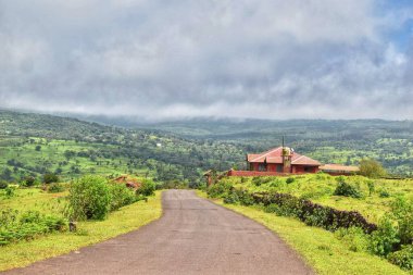 Sade bir kırsal yol yemyeşil tepeler boyunca esiyor, Satara, Maharashtra, Hindistan 'da bulutlu gökyüzünün altında kırmızı tuğlalı bir eve çıkıyor. Barışçıl kırsal cazibe canlı yeşillik ve açık manzaralarla gelişir..