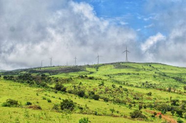 Devasa yeşil tepelerde Satara, Maharashtra 'da bulutlu mavi gökyüzüne karşı modern rüzgar türbinleri bulunur. Huzurlu manzara doğa ve yenilenebilir enerjinin birleşimini vurgular.