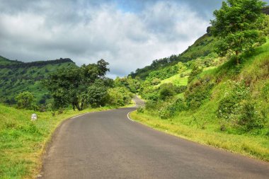 Satara, Maharashtra 'da yemyeşil tepelerden geçen kıvrımlı bir yol, yemyeşil ve bulutlu bir gökyüzü altında sakin manzaralarla çevrilidir..
