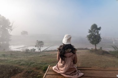 Araku Vadisi, Andhra Pradesh 'te sisli bir sabah manzarasına bakan ahşap bir platformda oturan beyaz bereli ve ceketli bir kadın..
