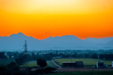 Jawai, Rajasthan 'ın sisli dağlarının üzerinde nefes kesici bir gün doğumu, altın renkleri tarlaları, kırsal evleri aydınlatıyor, ve dağınık ağaçları.