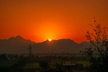 Hindistan, Rajasthan 'daki engebeli Jawai tepelerini aydınlatan turuncu gün batımı. Siluetli ağaçlar, uzak dağlar ve sakin bir kırsal alan huzur dolu manzarayı tamamlıyor..