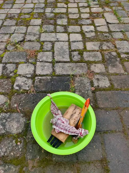 Üzerinde çeşitli temizlik ve boya aletleri bulunan canlı yeşil bir leğen yıpranmış taş kaldırımların üzerinde duruyor. Bu da açık hava bakımı ya da el işi olduğunu gösteriyor..