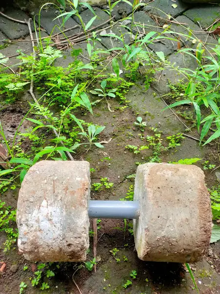 Yakın plan çekimde, beton bloklar ve borudan yapılmış ve yeşil otlarla kaplı taştan bir patikada dinlenen ev yapımı bir dambıl görülüyor. Bu resim yeterliliği ve fitness için temel yaklaşımı somutlaştırır..