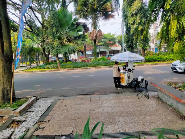 Mobil bir kahve satıcısı, Malang 'da kaldırımda beyaz şemsiyeli bir el arabası kullanıyor ve ağaçlı bir cadde boyunca müşterilere içki servisi yapıyor. Doğu Java. 22 Haziran 2025