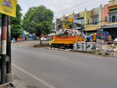 Bu görüntü kentsel atık yönetiminin katıksız gerçekliğini yansıtıyor, sokakta daha fazla atıklarla çevrili canlı sarı çöp kamyonu ile birlikte. Malang, Doğu Java. 24 Haziran 2025