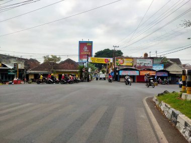 Bu fotoğraf, Endonezya 'nın Malang şehrinde kalabalık bir kavşağı yakalıyor, motosikletler park halinde ve sürüş halinde, yerel dükkanlar ve reklam panoları geniş bir gökyüzünün altında, tipik şehir yaşamını gösteriyor. Doğu Java. 24 Haziran 2025