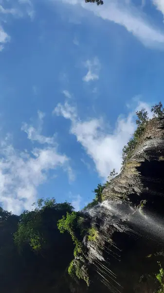 Yukarıya bakan bir perspektif kayalık bir uçurumun üzerinden çağlayan bir şelale yakalar, su çizgileri dağınık beyaz bulutlarla dolu parlak mavi bir gökyüzüne karşı görünür. Şelalenin doğal gücünü ve güzelliğini vurgular..
