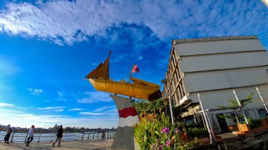 21 Haziran 2025 'te Pontianak' taki Rıhtım Kenti 'nin kıyısındaki Altın Tekne Heykeli' nin önünde atmosferin tadını çıkaran üç kişi yürüyor.