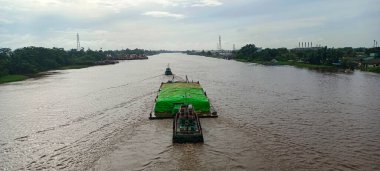 Arkasından küçük bir römorkör tarafından itilen, büyük, düz güverte mavnası, ağır yüklü yeşil bir branda ile kaplı kargo.