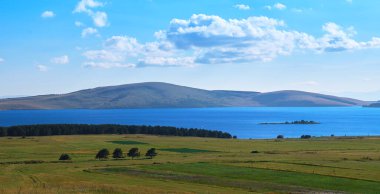 Bereketli yeşil tarlalar güneşli güneşin altında kayan tepelerle çevrili sakin sularla buluşur.