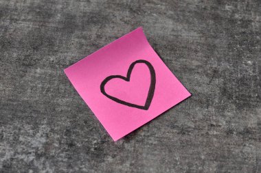 Üzerinde kalp çizimi olan pembe yapışkan bir not gri bir yüzeyde duruyor.