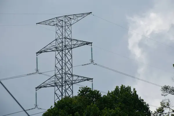 Yüksek voltajlı elektrik direği Bulutlu Gökyüzüne karşı Yeşil Ağaçlarla