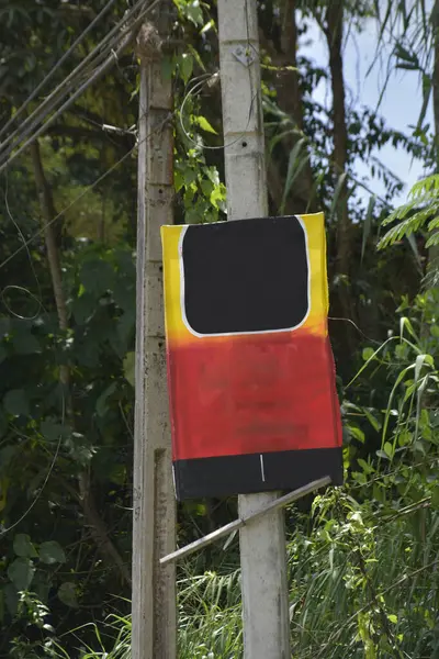 Kırsal beton elektrik direğine tutturulmuş, siyah dikdörtgen bir üst ve alt şeridi olan koyu kırmızı ve sarı kısımları içeren merak uyandırıcı, elle boyanmış soyut bir tabela. Ağaçların ve yaprakların yemyeşil doğal arka planına karşı kuruludur.
