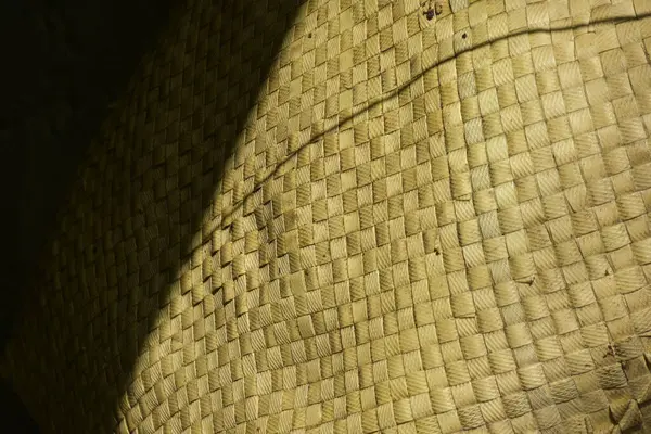 Yakın plan, yüksek açılı doğal, altın rengi dokuma dokuma dokusunun muhtemelen bir hasırdan, sepetten ya da geleneksel el işçiliğinden. Güneş ışığı, karmaşık kare dokuma boyunca keskin çapraz gölgeler oluşturur. Çiğ, organik güzelliğini ve dokunsal kuasını vurgular.