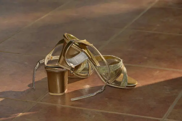 Parıltı ve metalik kayışlarla süslenmiş göz kamaştırıcı yüksek topuklu sandaletler, kahverengi fayanslı zeminde gelişigüzel duruyor. Onların yanında, bir olay ya da kutlama hissine eşleşen altın renkli bir kupa eklenir. Bu görüntü moda temalarını çağrıştırıyor.,