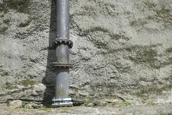 Görünür flanşlar ve cıvatalarla korunan sağlam bir endüstriyel metal boru, kısmen yosun veya renk bozulmasıyla kaplı sert, desenli beton bir duvara dikey olarak uzanır. Resim altyapı, kamu hizmetleri, hammadde temalarını vurguluyor,