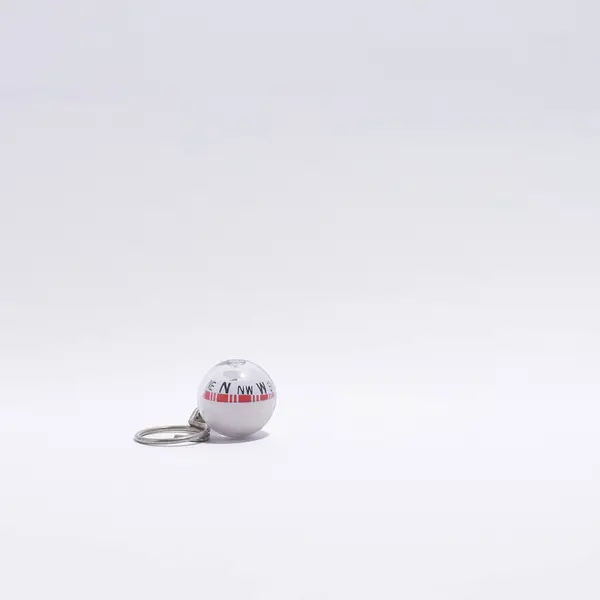 Küçük, şeffaf küresel bir pusula anahtarlığı, beyaz bir taban ile görünür kardinal yönler ve metalik bir halka, belirgin beyaz bir zemin üzerinde durmaktadır