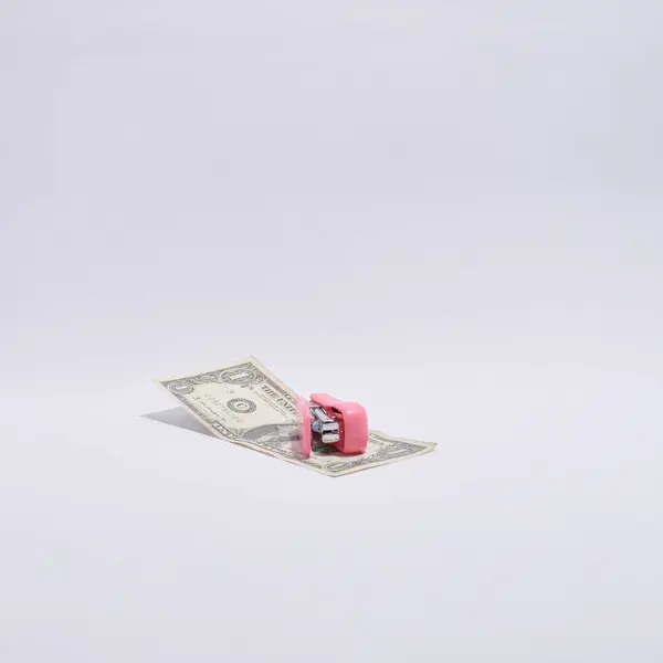 Tek bir Amerikan doları kısmen katlanır ve parlak pembe bir zımba tarafından tutulur, temiz, kıvrımlı beyaz bir arka planda sunulur.