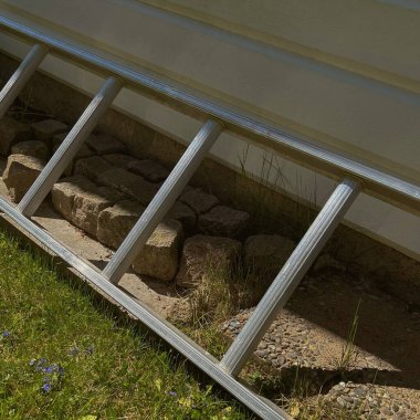 Beyaz saray duvarının yakınındaki çimlere ve taşlara yerleştirilmiş metal bir merdivenin ayrıntılı yan görüntüsü. Onarım ve bakım temaları, donanım görselleri veya bahçe aracı tasarımı için mükemmel.