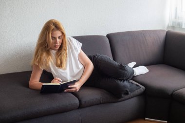 Evdeki kanepede kitap okuyan genç bir kadın.