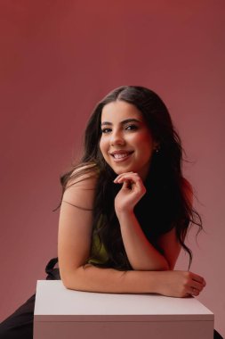 Uzun siyah saçlı genç bir kadın usulca gülümsüyor. Beyaz bir bluz giyer ve kameraya tarafsız bir arkaplanda bakar. Kendine güveni ve dostluğu yayar..