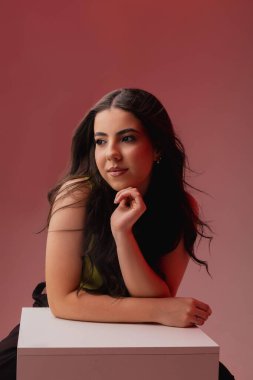 Uzun siyah saçlı genç bir kadın usulca gülümsüyor. Beyaz bir bluz giyer ve kameraya tarafsız bir arkaplanda bakar. Kendine güveni ve dostluğu yayar..