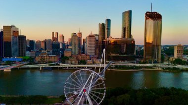 Günbatımında Queensland, Avustralya 'da Brisbane CBD' nin hava görüntüsünde parlayan şehir silueti, yüksek binalar, Brisbane 'nin ikonik tekerleği ve Brisbane Nehri yer alıyor. Altın saat boyunca yakalanan bu resim canlı kenti vurguluyor.
