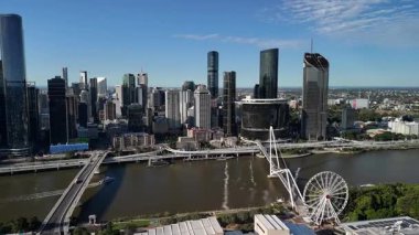 Queensland, Avustralya 'daki Brisbane City' nin gündüz hava görüntüsü. Görüntü, kentin modern silueti, Brisbane Nehri, önemli köprüler ve parlak mavi gökyüzü altında yükselen ticari binaları gözler önüne seriyor. Merkezi ticaretin net ve geniş bir görüntüsü