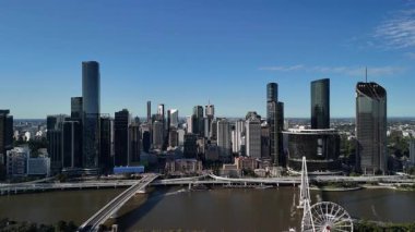 Queensland, Avustralya 'daki Brisbane City' nin gündüz hava görüntüsü. Görüntü, kentin modern silueti, Brisbane Nehri, önemli köprüler ve parlak mavi gökyüzü altında yükselen ticari binaları gözler önüne seriyor. Merkezi ticaretin net ve geniş bir görüntüsü