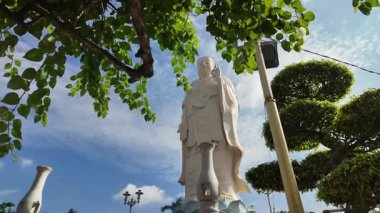 Vietnam 'daki My Tho' daki Vinh Trang Pagoda 'da çekilen videoda beyaz Arhat heykelleri ve dalgalanan Budist bayraklarıyla çevrili görkemli gülümseyen Buda heykeli görülüyor. Hareket eden bulutlar ve buradan geçen ziyaretçilerle çarpıcı bir gökyüzünün altında.