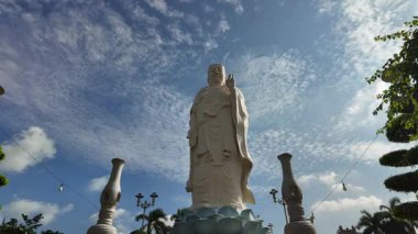 Vietnam 'daki My Tho' daki Vinh Trang Pagoda 'da çekilen videoda beyaz Arhat heykelleri ve dalgalanan Budist bayraklarıyla çevrili görkemli gülümseyen Buda heykeli görülüyor. Hareket eden bulutlar ve buradan geçen ziyaretçilerle çarpıcı bir gökyüzünün altında.