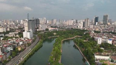 Hanoi, Vietnam 'daki Thu Le Lake' in 4K hava görüntüsü, su, yürüyüş yolları ve şehrin yüksek ufuk çizgisinin arka planında yer alan ağaçla kaplı huzurlu yeşil bir vahayı gözler önüne seriyor. Bu manzaralı kentsel göl,
