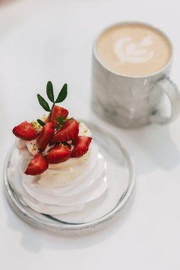 Zarif bir kafe anı: üzerinde taze çilek parçaları, şam fıstığı kırıntıları ve yeşillik bulunan özel bir pavlova mermer bir tabakta, parlak beyaz bir masa üzerinde yaprak sanatı ile kremalı sütlü bir latte ile birlikte oturur.