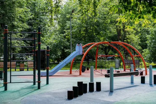 Yapraklı parka yerleştirilmiş renkli bir şehir oyun alanının geniş bir görüntüsü: Mavi kaydırak, turuncu kemer tırmanma ağı ve fitness çubukları güvenli lastik döşeme üzerinde duruyor, banklar ve yaz yeşiliyle çerçevelenmiş, rahatlamış aile eğlence alanını insanlar olmadan yakalama