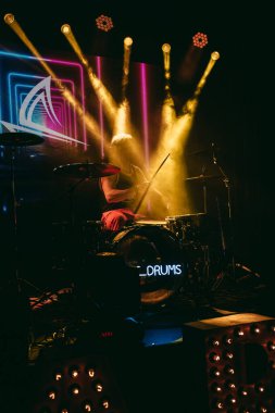 Enerjik bir davulcu canlı performans sergiliyor. Etrafı renkli ışıklar ve canlı bir atmosferle çevrili.