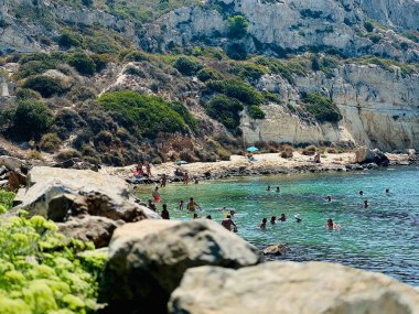 İtalya 'nın Sardinya kentindeki güneşli sahil sahnesinde insanlar turkuaz sularda yüzüyor ve engebeli uçurumlarda dinleniyorlar. Canlı bir Akdeniz yaz havası. Tatil, yaşam tarzı ve seyahat promosyonları için mükemmel..