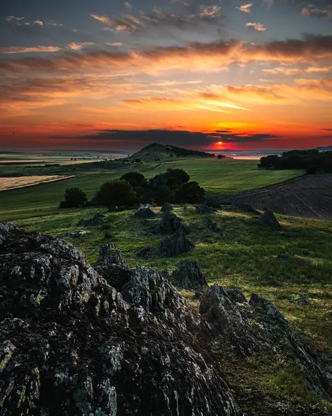 Nefes kesen bir günbatımı, engebeli bir manzaranın üzerinde turuncu ve kırmızı renkler halinde gökyüzünü boyar. Ön plandaki antik kaya oluşumları gözü yuvarlanan yeşil tepelere yönlendirir..