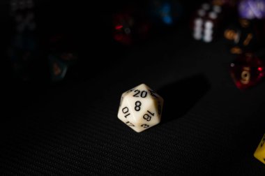 Tek bir beyaz 20 kenarlı zar (D20), renkli ve bulanık RPG zarlarının arka planına karşı keskin bir odak noktasında duruyor. Dramatik ışıklandırma ana konuyu ve önemini vurguluyor..