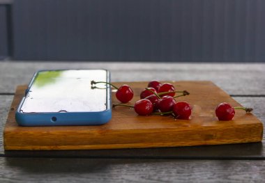 Telefon, siyah bir masanın üzerinde kırmızı bir kirazın yanında duruyor.