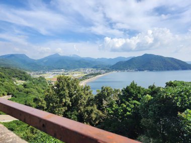 Bereketli yeşil dağlar ve sakin mavi deniz arasında kurulmuş huzurlu bir kıyı köyünün manzaralı bir manzarası. Aydınlık ve açık bir günde çekilen bu görüntü, kırsal Japonya 'nın sessiz güzelliğini ve doğa ile insan yerleşimi arasındaki uyumu yansıtıyor.. 