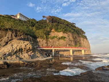 Turistler Japonya 'da deniz kenarındaki bir uçuruma inşa edilmiş çarpıcı kırmızı raylı bir yol boyunca yürürler. Tepedeki gür yeşillik, dokulu kaya oluşumları ve batan güneşin altın ışığı huzurlu ve dramatik bir atmosfer yaratır..