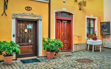 Ahşap kapılı renkli binalar, saksı bitkileri ve çiçeklerle dolu bir masa. Sahne ilginç ve davetkar bir atmosfer yakalıyor..