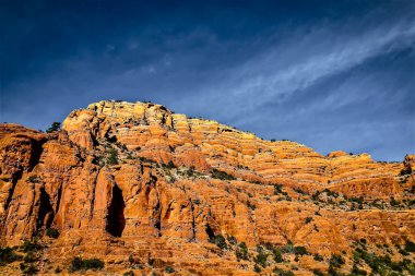 Mavi gökyüzünün altında parlak turuncu ve kırmızı renkleri olan kayalık bir uçurumun çarpıcı manzarası. Kayaların dokusu belirgin, çöl manzaralarına özgü katmanlar ve oluşumlar sergiliyor..