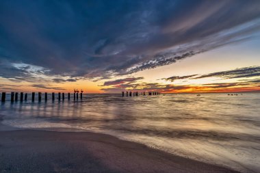 Suya uzanan siluetli ahşap sütunlarla sakin bir denizin üzerinde sakin bir günbatımı. Gökyüzü dramatik bulutlarla doludur. Turuncu, pembe ve mavi tonlarında. Nazik dalgaları yansıtır..