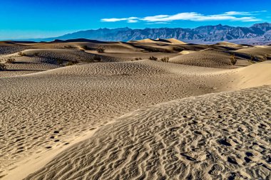Uçsuz bucaksız bir çöl manzarasında açık mavi bir gökyüzünün altında kum tepeleri yuvarlanıyor. Uzak dağlar çarpıcı bir zemin oluşturur, sahnenin doğal güzelliğini arttırır..