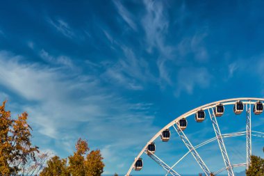 Parlak bir dönme dolap açık mavi gökyüzüne karşı ince bulutlu, sonbahar yapraklarıyla çevrili..