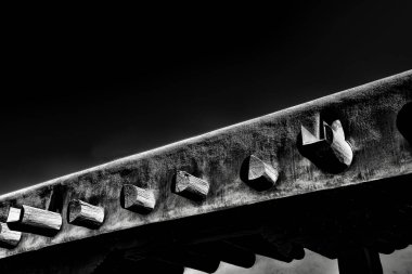 Siyah ve beyaza yakalanmış, ahşap saplı, çıkıntılı ahşap bir kirişin yakın çekimi. Görüntü, ışık ve gölge arasındaki zıtlığı vurguluyor, işçiliği vurguluyor.