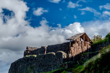 Bulutlu dramatik bir gökyüzünün altındaki bir yamaçta tarihi bir taş yapı. Binanın sazdan bir çatısı var ve çevresi yemyeşil, antik mimari sergileniyor..
