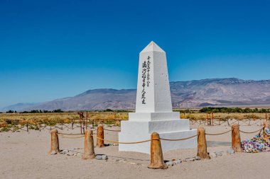 Üzerinde yazıtlar olan uzun beyaz bir anıt açık mavi gökyüzünün altında çorak bir arazide duruyor. Anıtın etrafında ahşap direkler ve arka planda dağlar bulunan kayalık bir sınır bulunuyor..